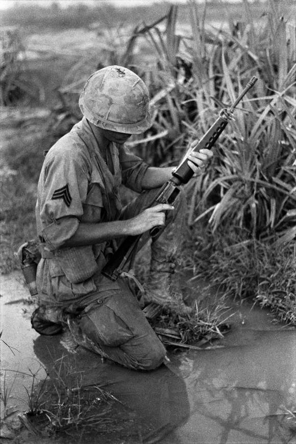 Um sargento ajoelha-se no chão molhado e verifica o seu M16. Nome, data e local desconhecido.