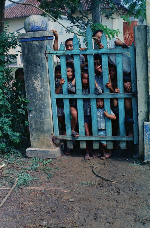 Crianças vietnamitas observam através de um portão a câmera de Haughey. Nomes, data e local desconhecidos.